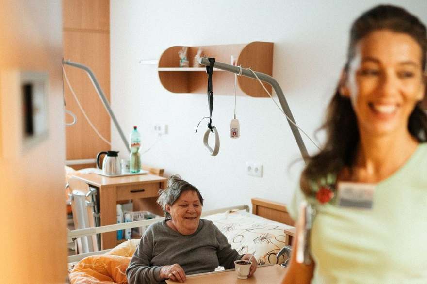 Pflegekraft betreut lächelnd eine ältere Frau in einem Pflegezimmer, Beispiel für Altenpflege Jobs